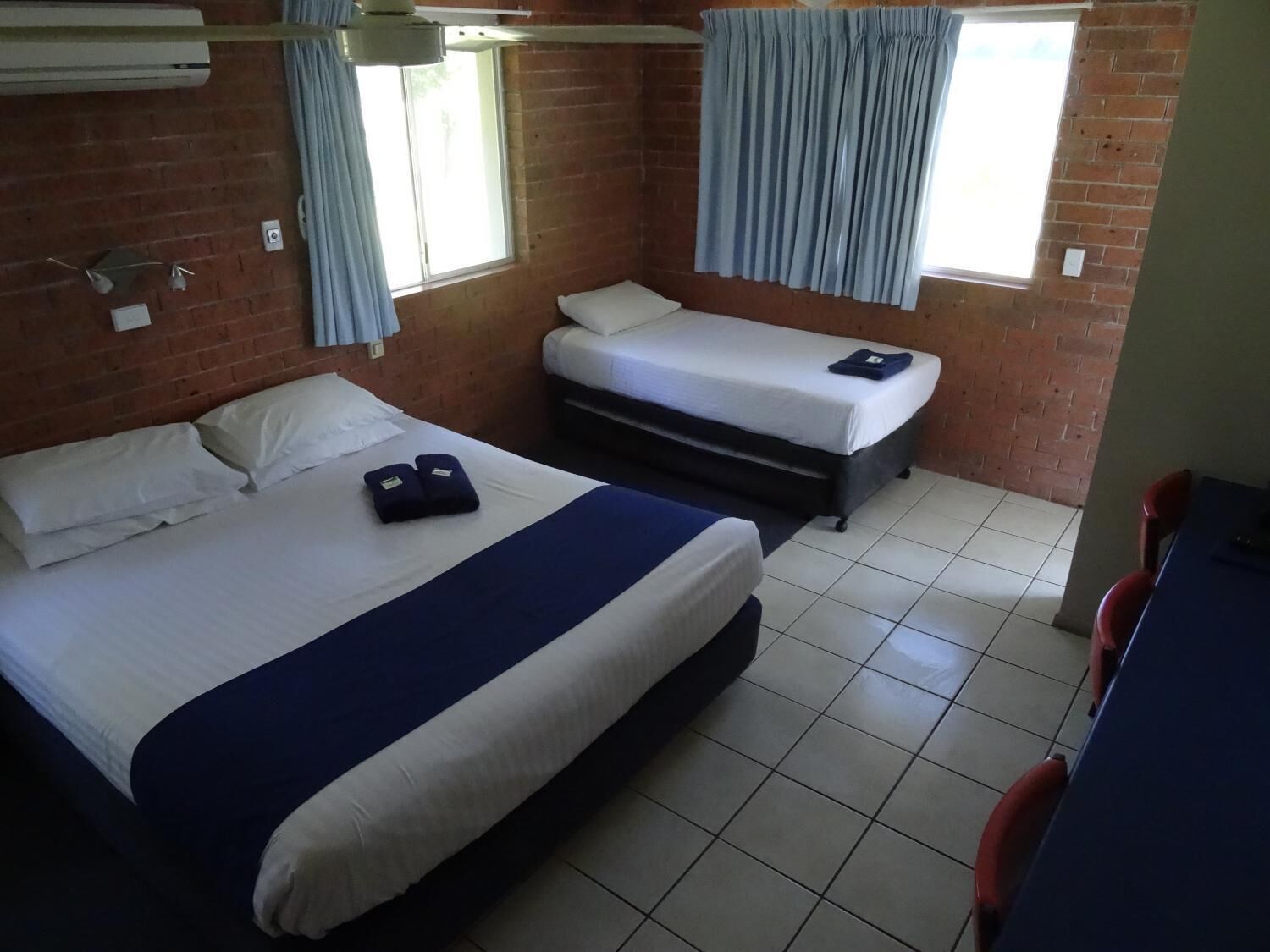 A hotel room with a large bed, a smaller bed, a desk, and a chair. There are two windows with curtains, and the room has a tiled floor.
