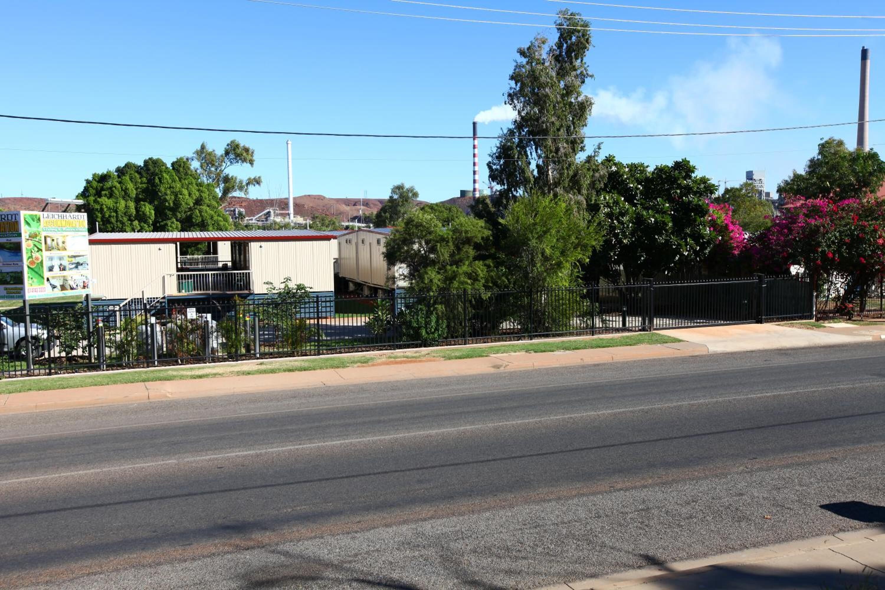 A street view with a fenced property, a building with a signboard, and trees.
