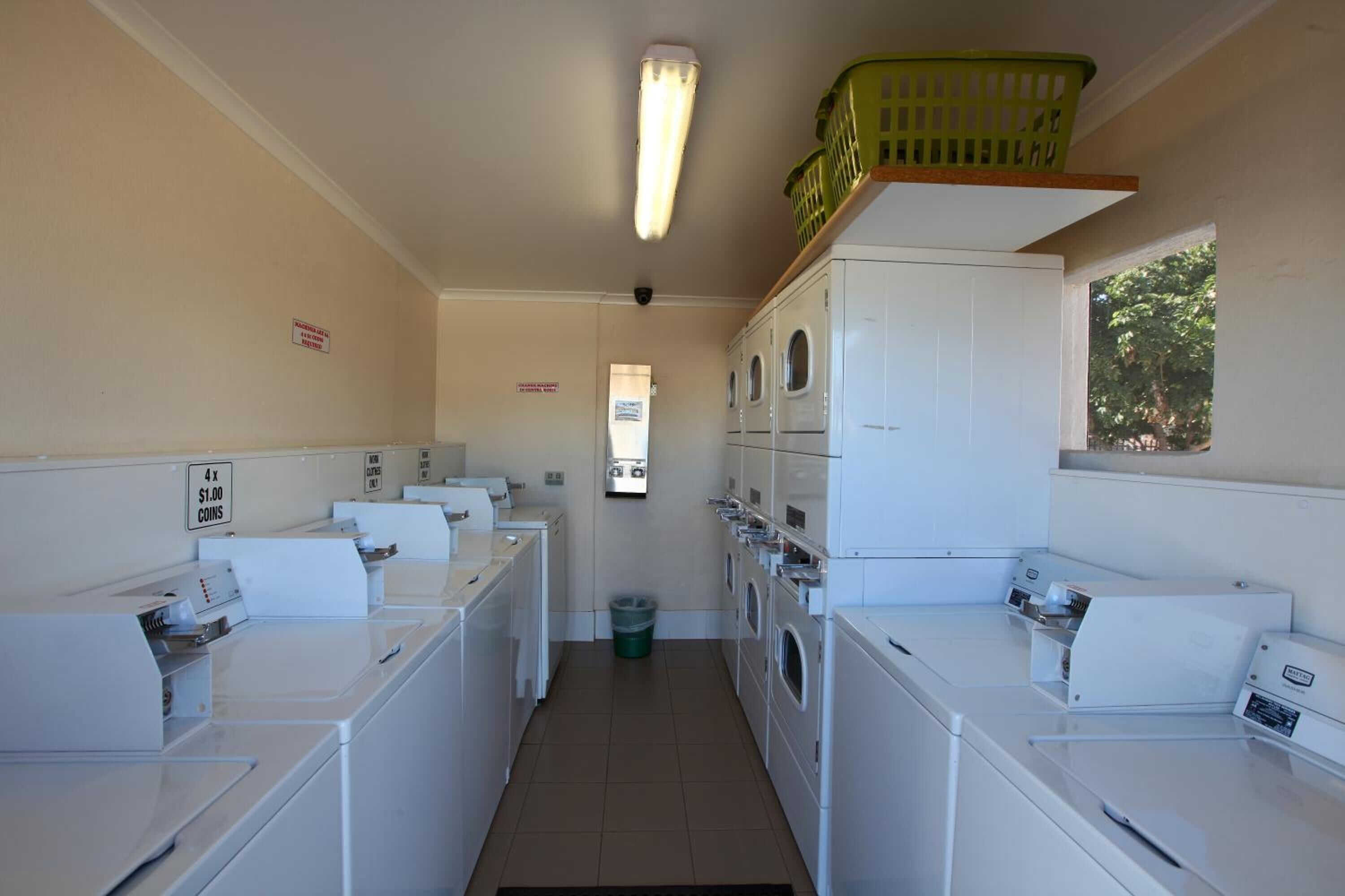A laundry room with multiple washing machines and dryers, a green basket on a shelf, and a sign indicating coin usage for laundry services.