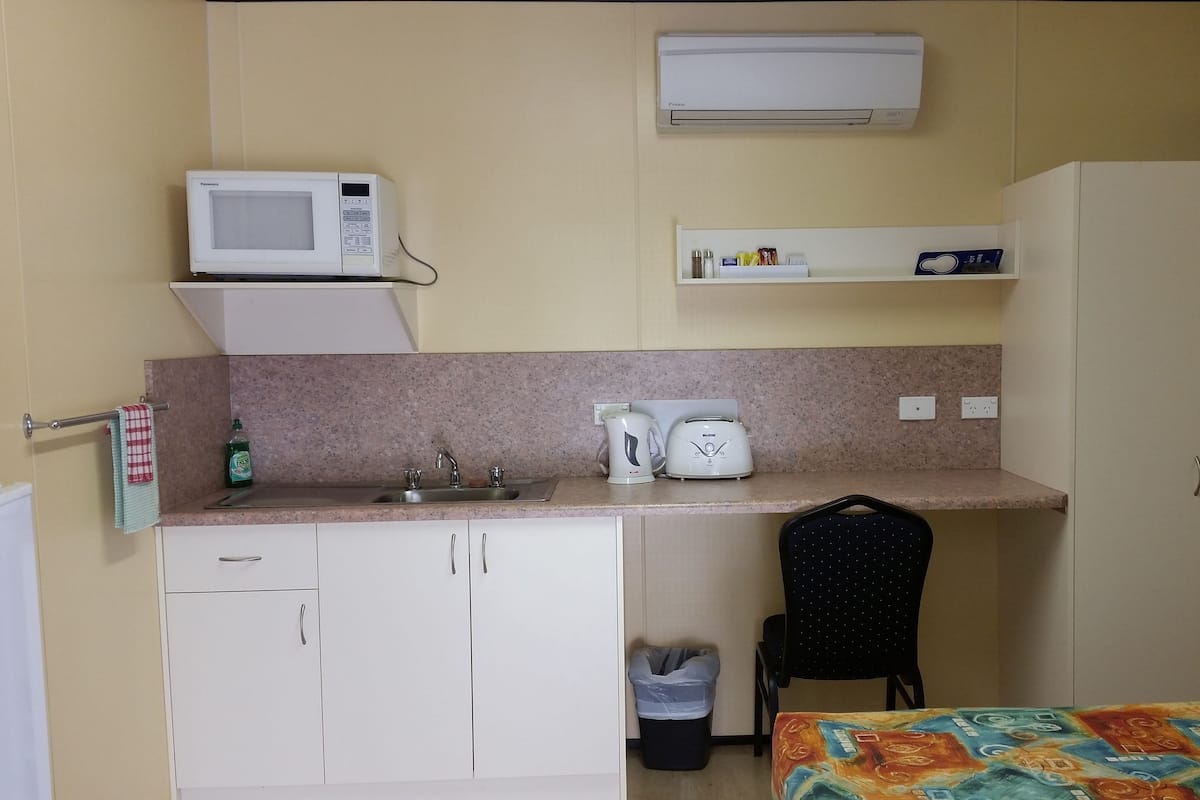 A compact kitchenette with a microwave, toaster, and sink. There is a small trash bin and a chair.