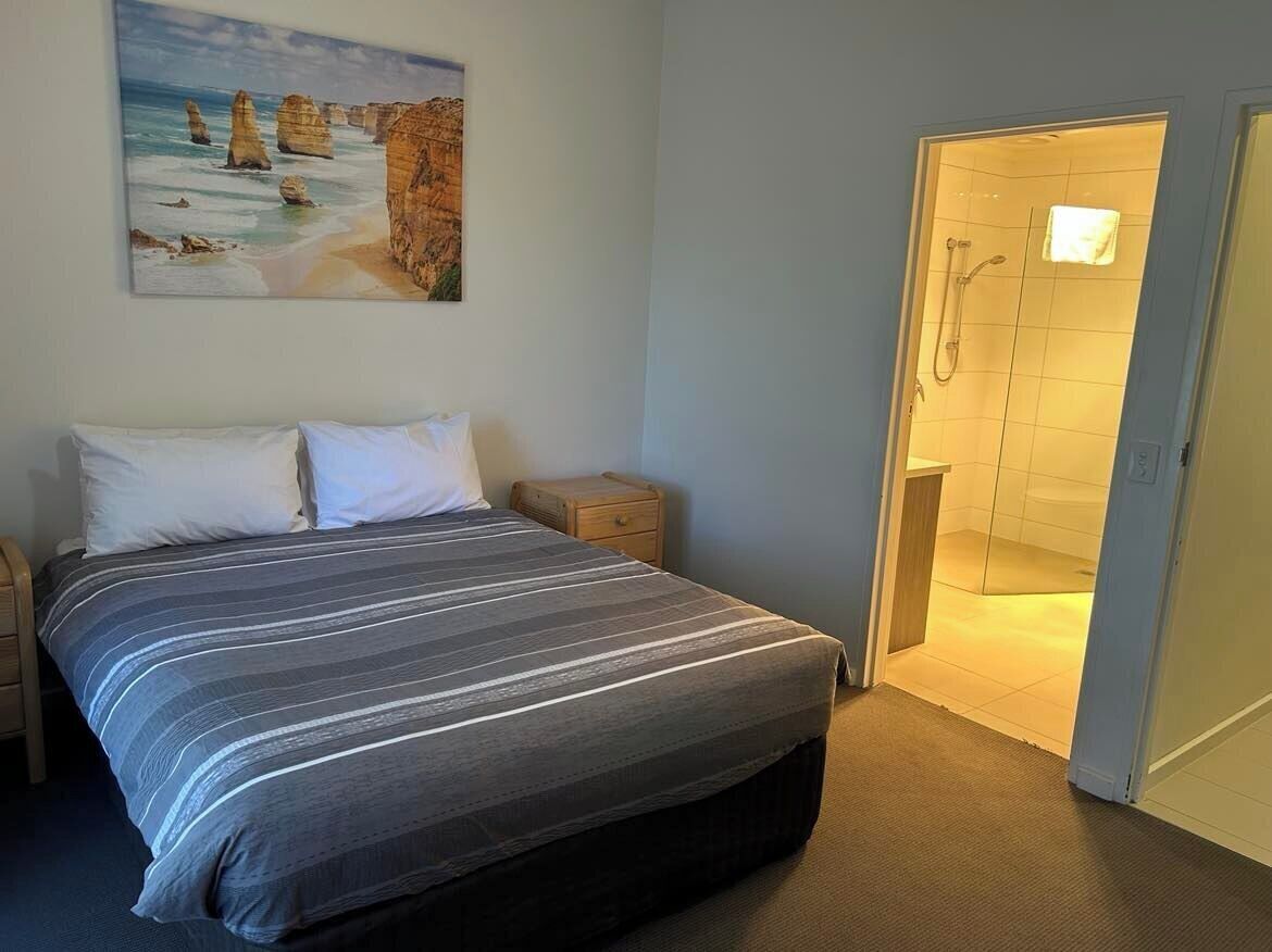 A bedroom with a bed, a nightstand, a painting of a coastal scene, and a shower area.