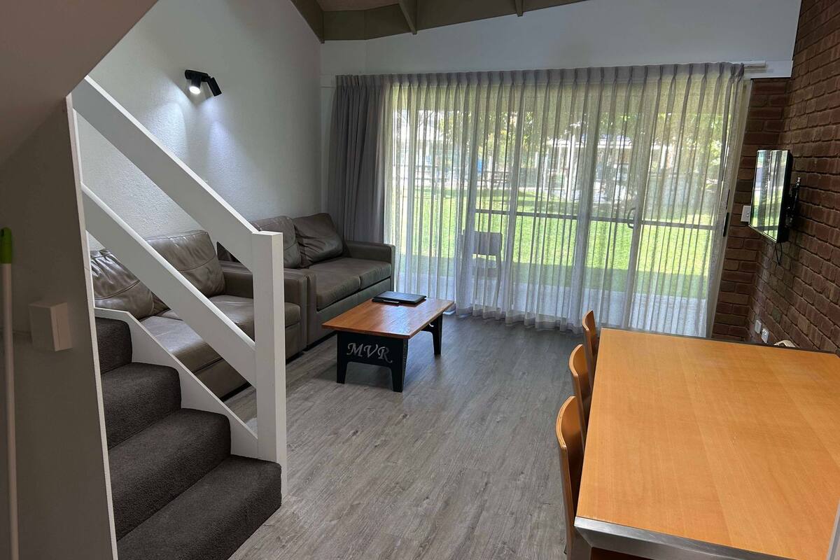 A modern living room with a staircase, a sofa set, a wooden coffee table, and a dining table with chairs.