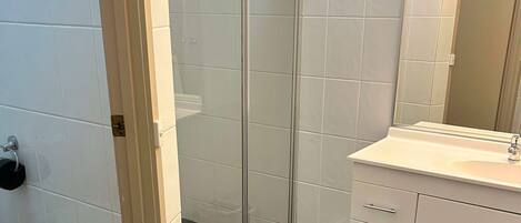 Un baño con cabina de ducha de vidrio, un lavabo blanco con espejo y piso de baldosas.