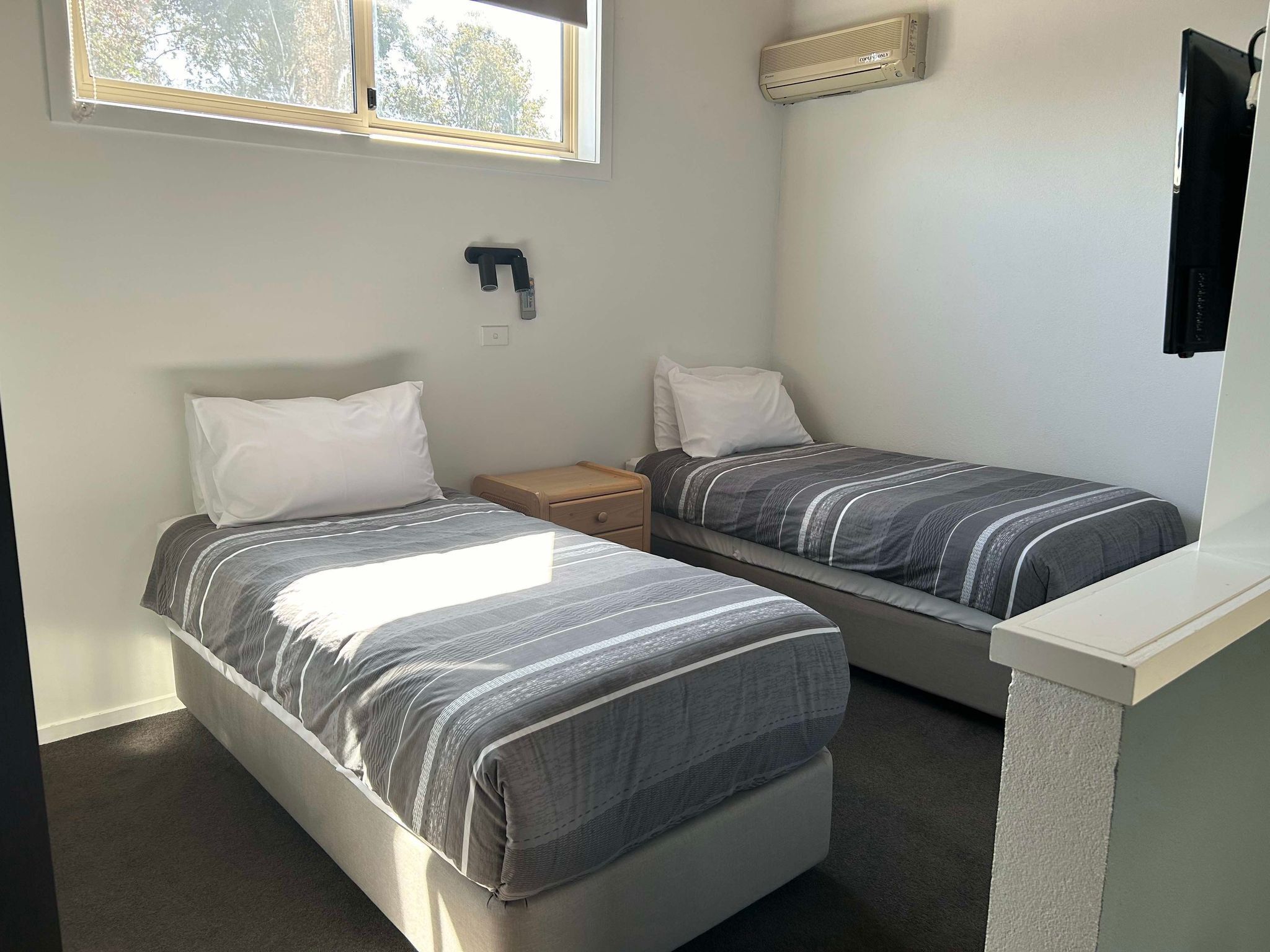 A small, clean room with two single beds, a nightstand, a wall-mounted heater, and a window with blinds.
