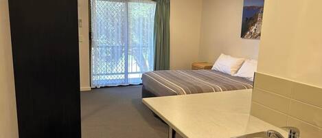 A hotel room with a bed, a small kitchenette with a sink, a ceiling fan, and a balcony with a view of the sea.
