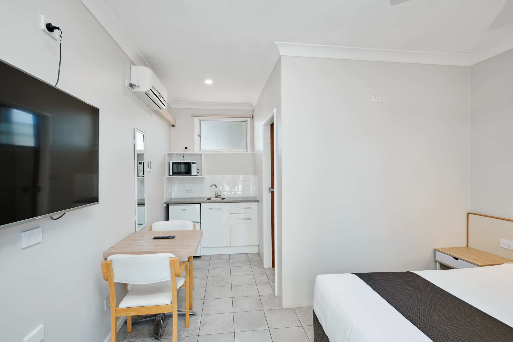 Habitación de hotel pequeña con cama, escritorio con silla, televisor en la pared, microondas y kitchenette.