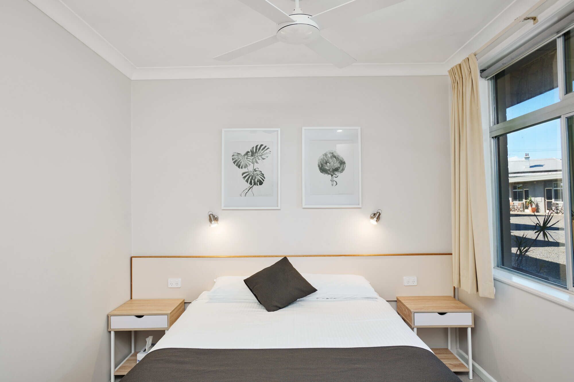 A bedroom with a bed, two bedside tables, a ceiling fan, and two framed botanical prints on the wall.