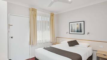 Une chambre d’hôtel comprenant un lit, un ventilateur de plafond, une fenêtre avec des rideaux, une table de chevet et une lampe fixée au mur.
