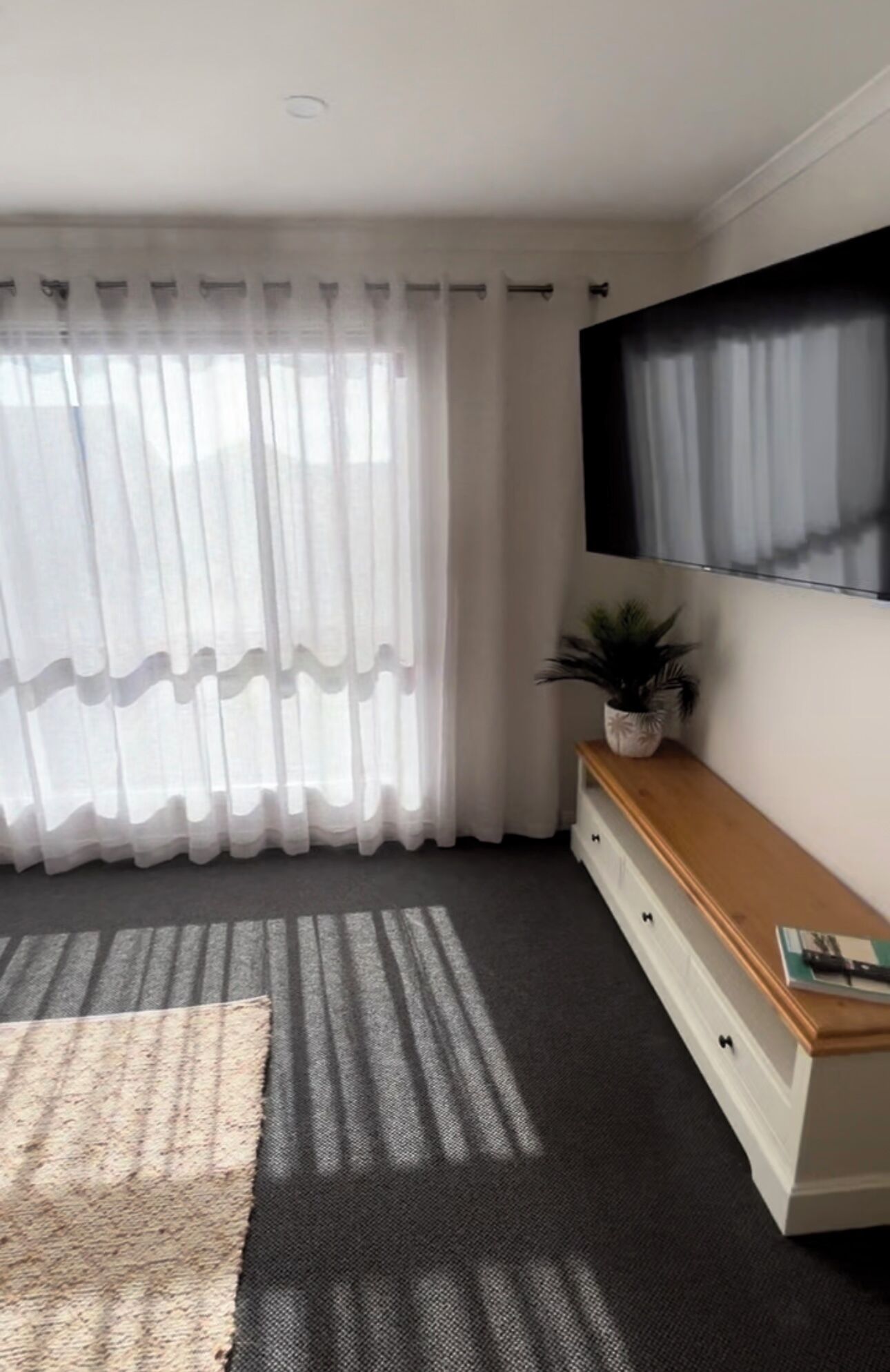 A modern living room with a large flat-screen TV mounted on the wall, a wooden TV stand with a plant, and a large window with sheer curtains.