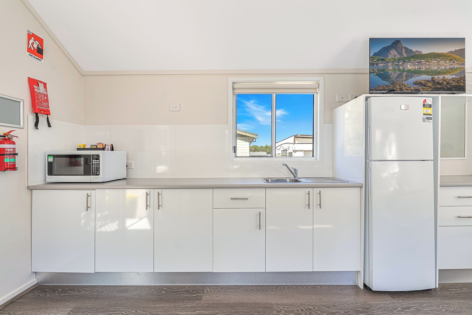 A kitchen with white cabinets, a microwave, a refrigerator, a window, and a fire extinguisher.