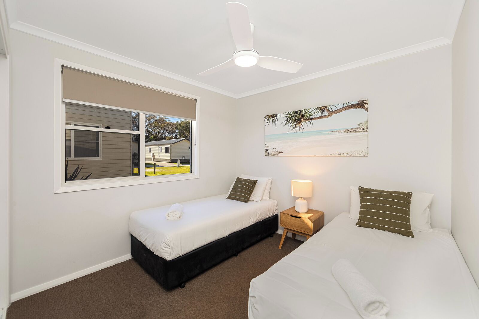 A bedroom with a single bed, a smaller bed, a nightstand with a lamp, a window with blinds, and a ceiling fan.