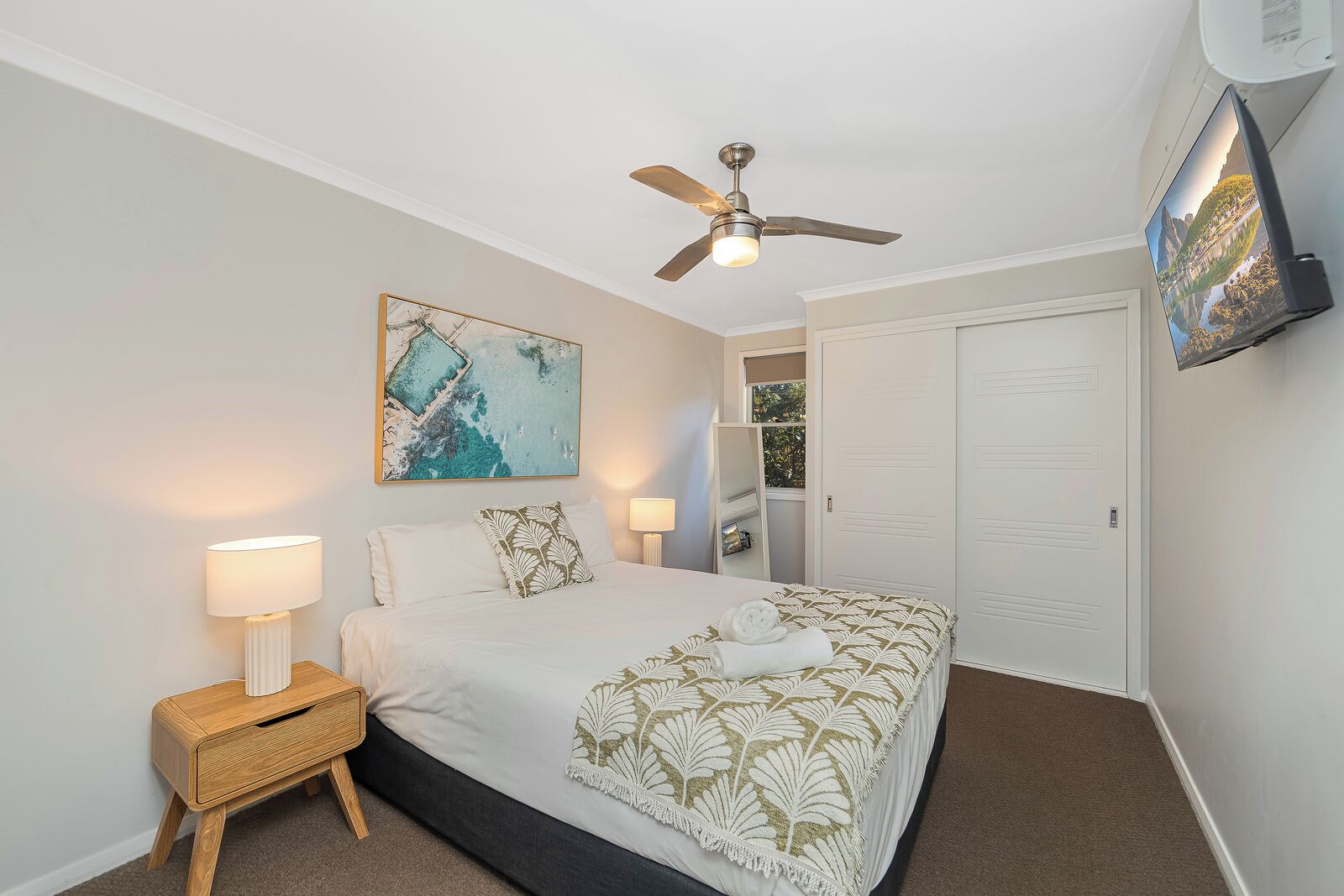 A hotel room with a bed, a ceiling fan, a painting, a television, and a wardrobe.