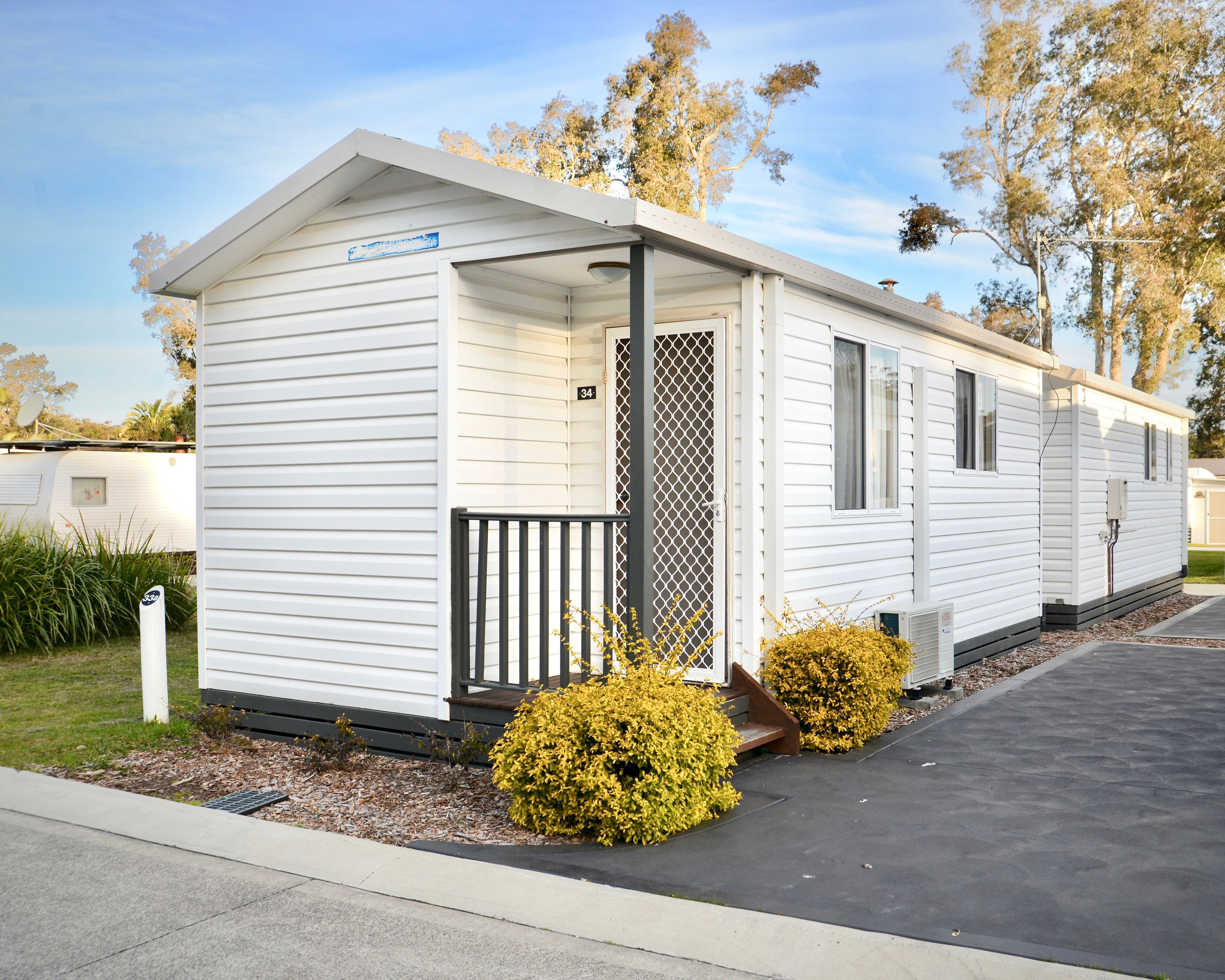 A single-story mobile home with a covered porch, a small front yard with yellow bushes, and a paved driveway.