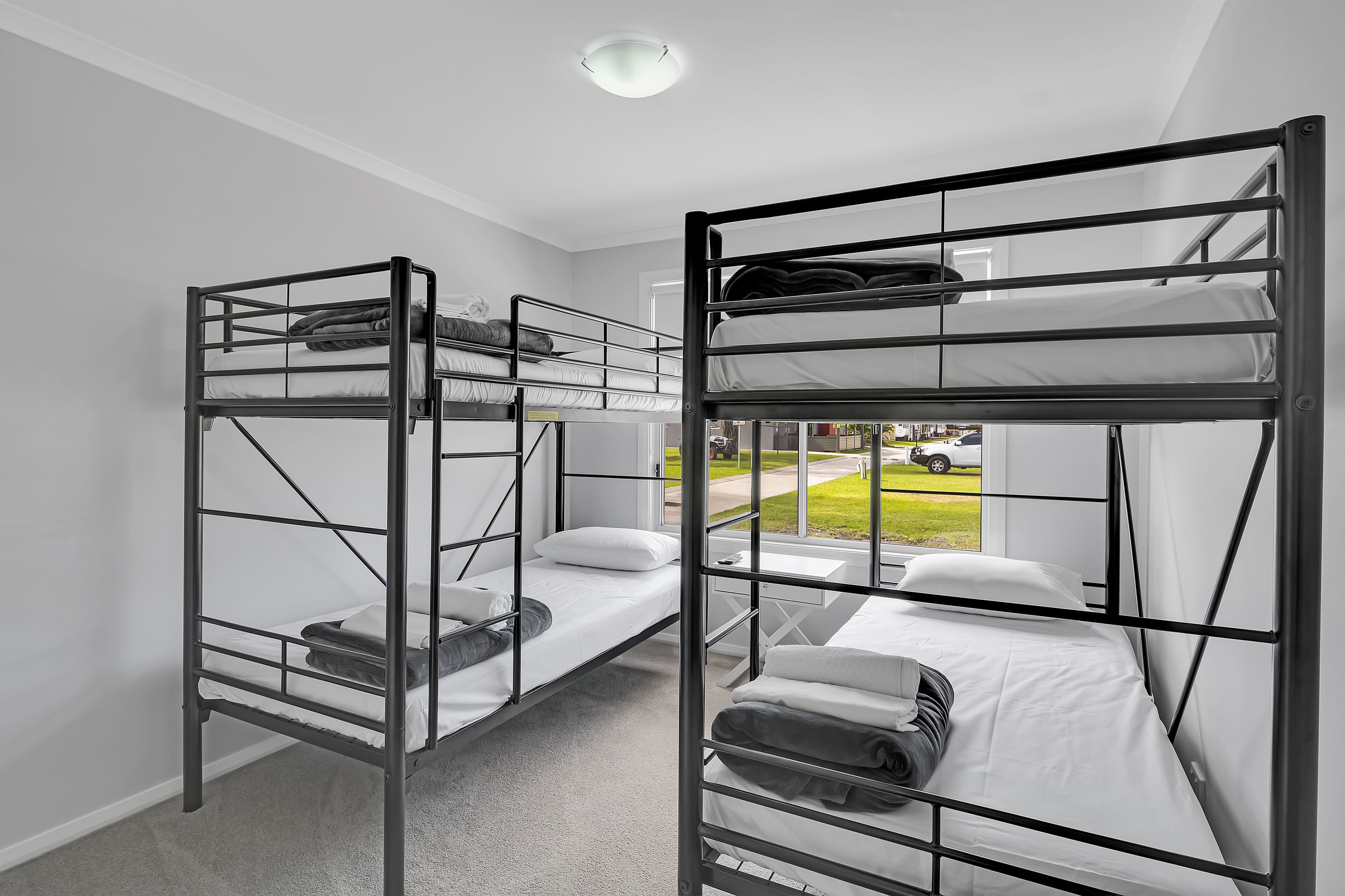 A room with two bunk beds, white walls, and a window with a view of a green lawn and parked cars.