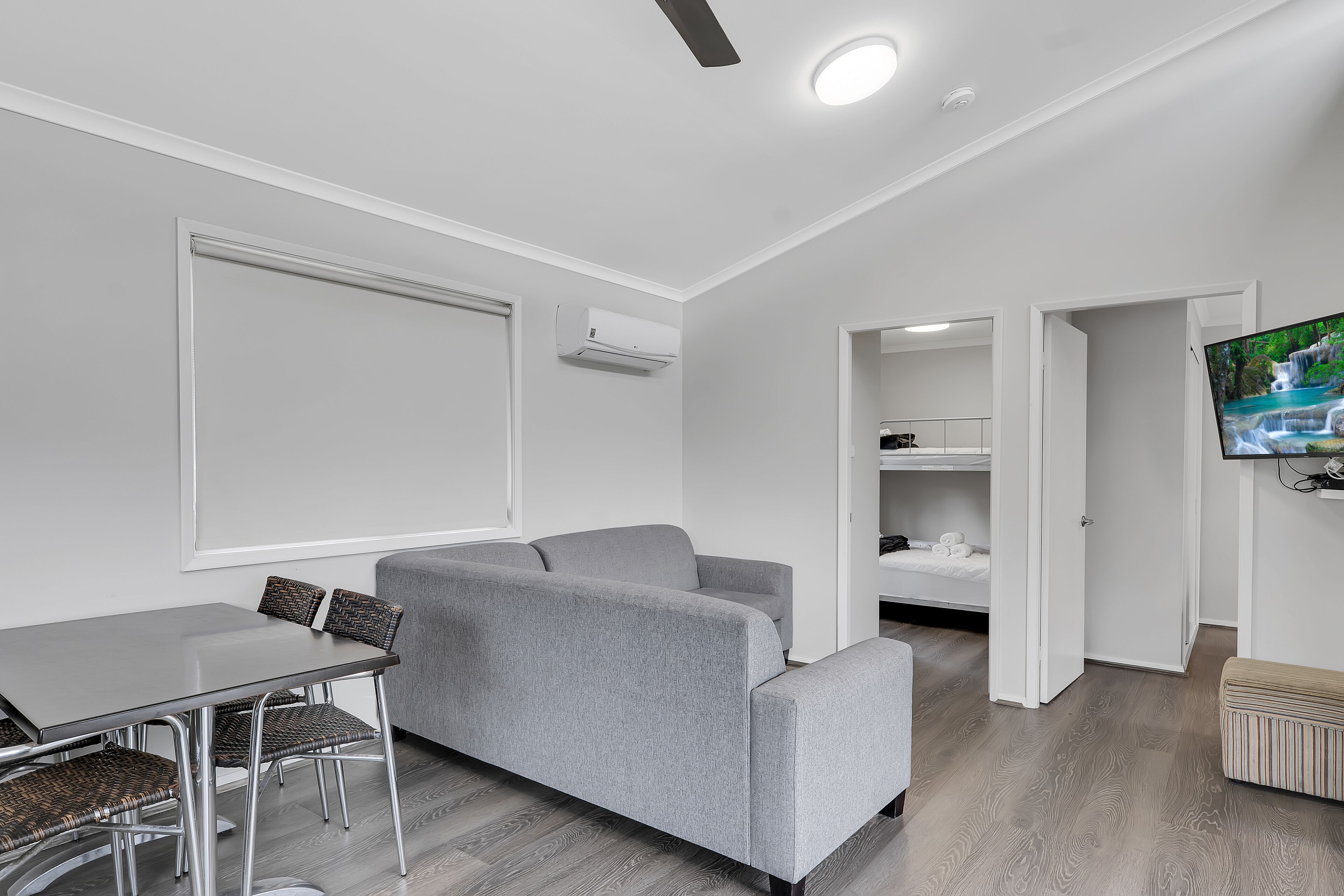 Een moderne woonkamer met een grijze bank, een eettafel met stoelen, een flatscreen televisie en een plafondventilator.