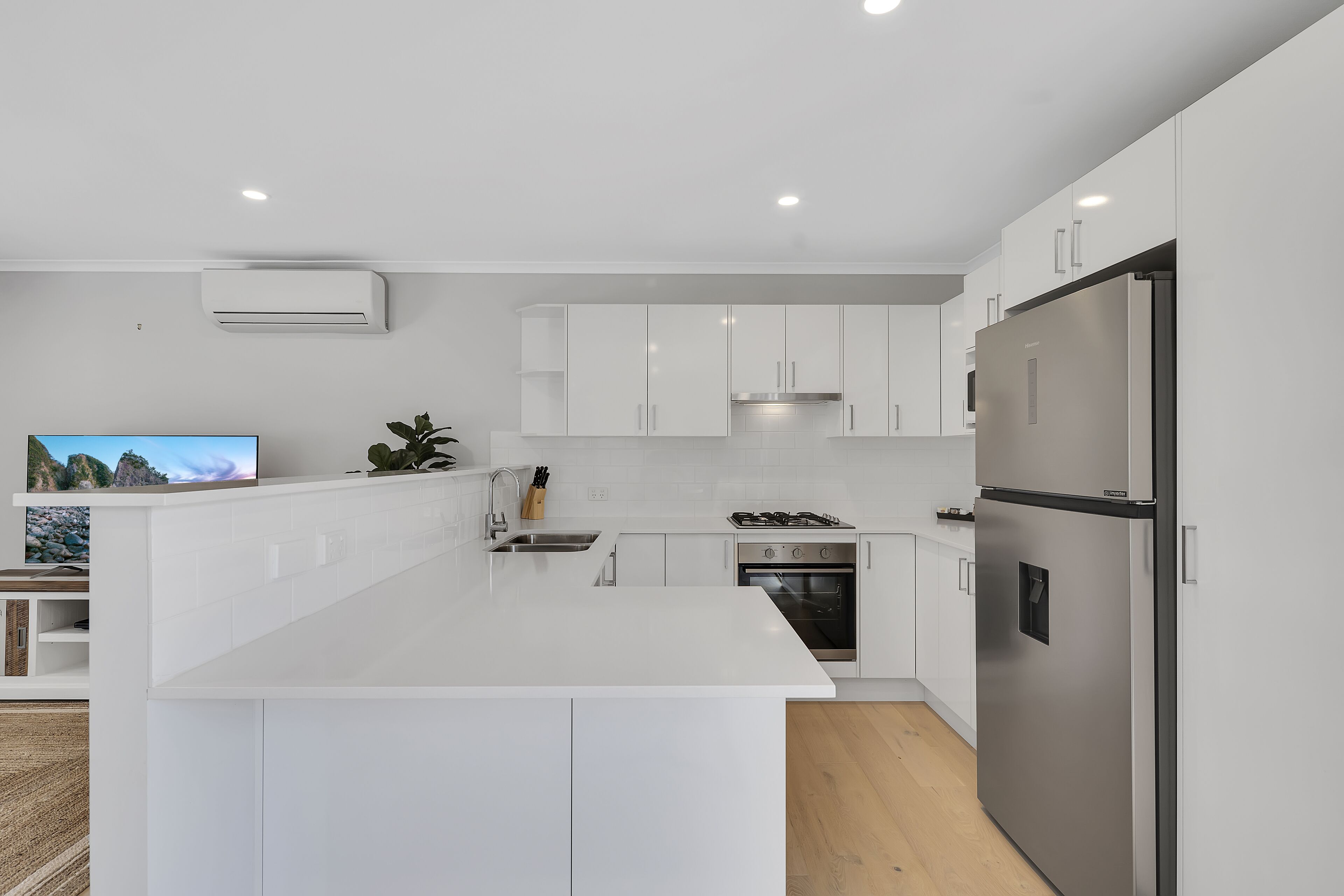 A modern kitchen with white cabinetry, a stainless steel refrigerator, a built-in oven, and a flat-screen TV on a counter.