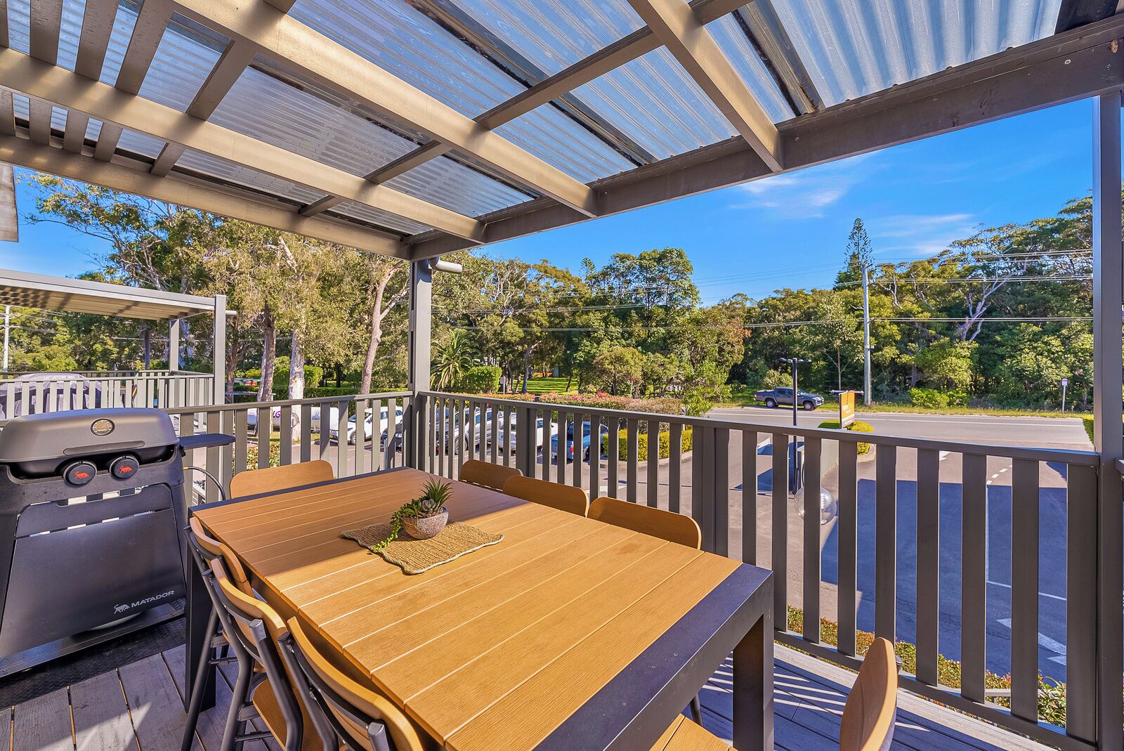 An outdoor patio with a grill, wooden table, and chairs, overlooking a road and greenery.