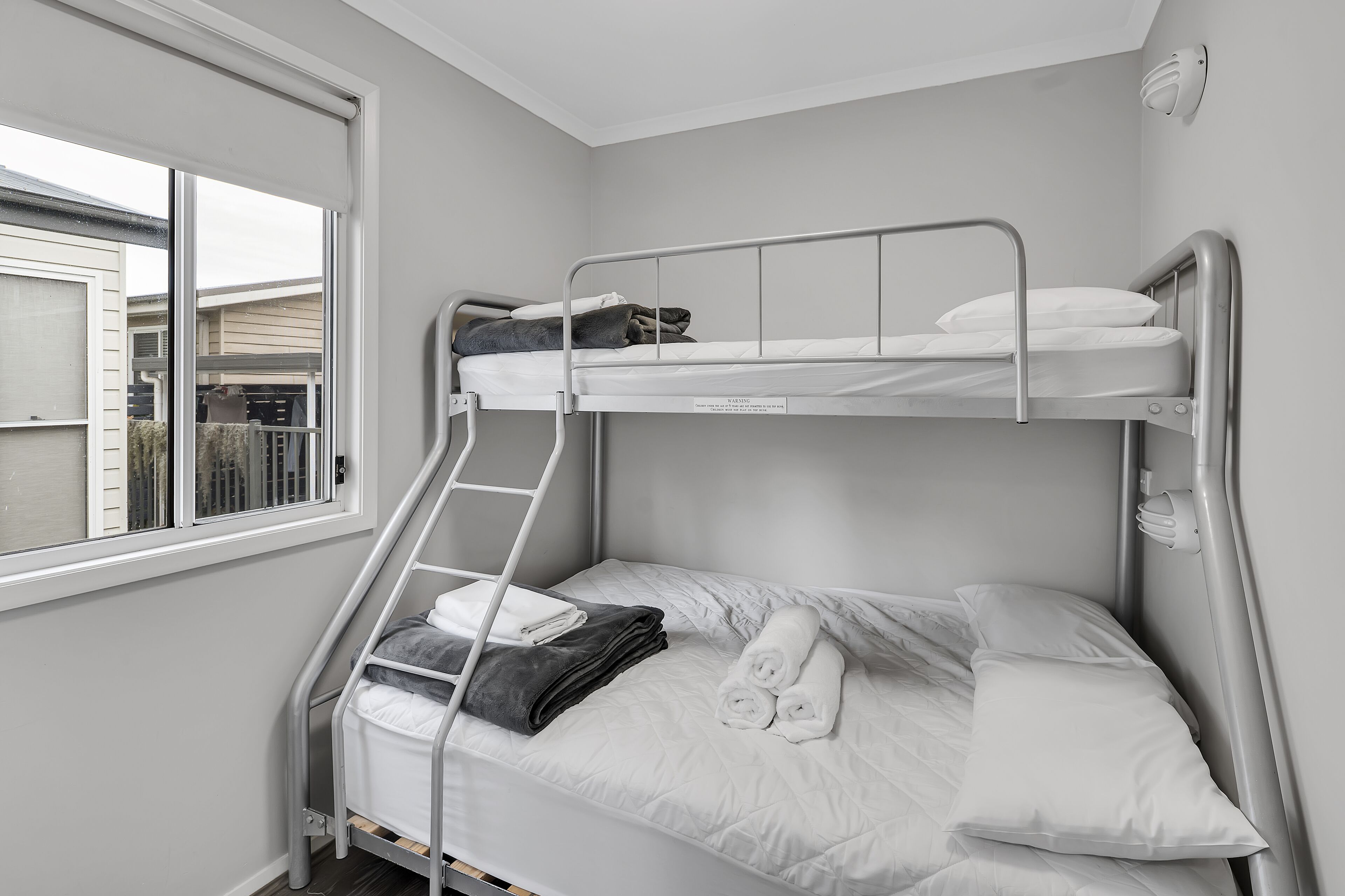 A bunk bed with white bedding and a ladder, a window with a view of a house, and a wall-mounted light fixture.