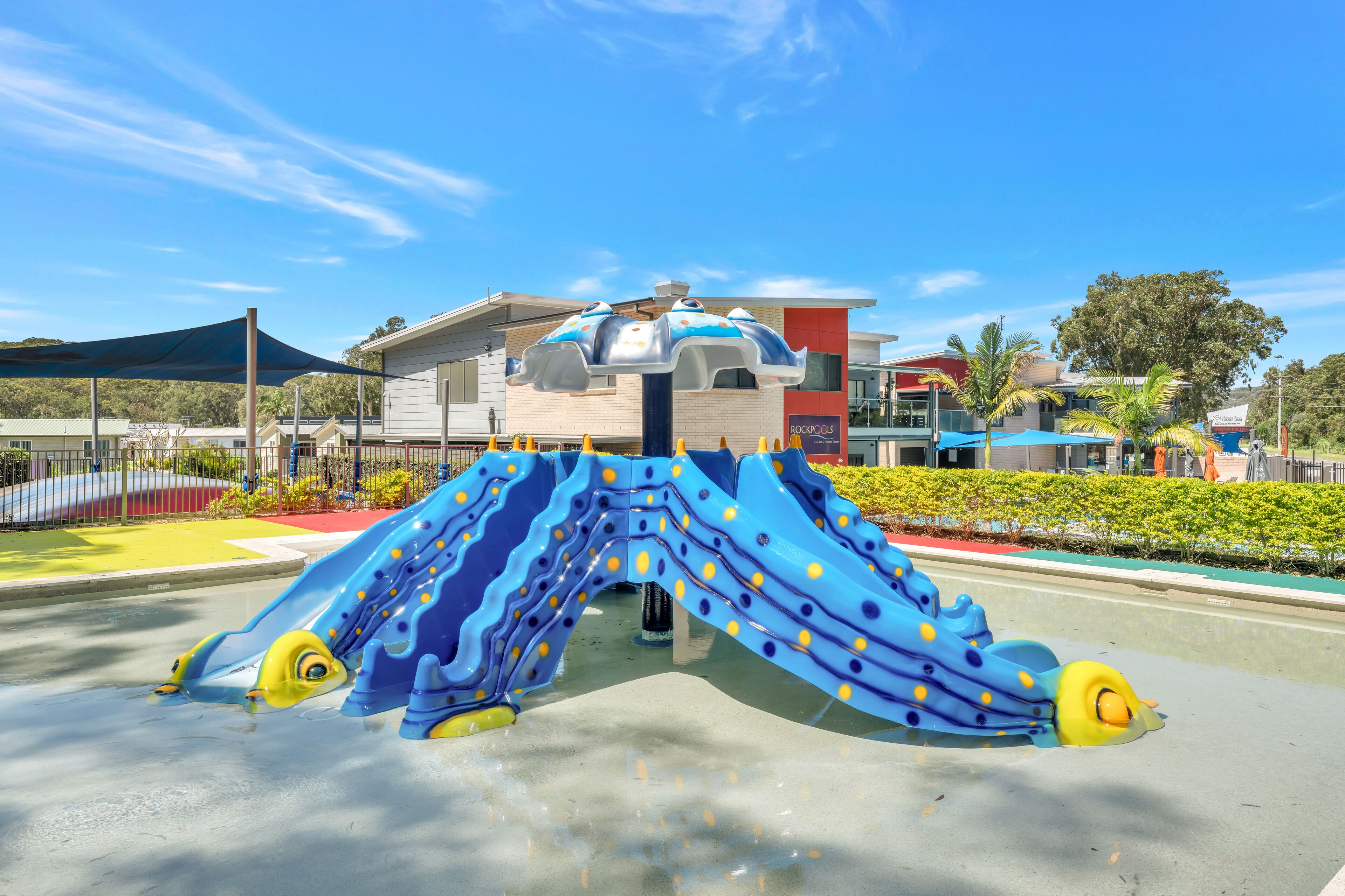 Une aire de jeux avec un toboggan bleu et des éléments décoratifs jaunes, entourée d’une clôture et de verdure.