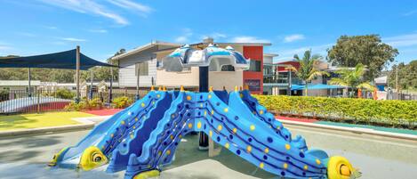 A playground with a blue slide and yellow accents, surrounded by a fence and greenery.