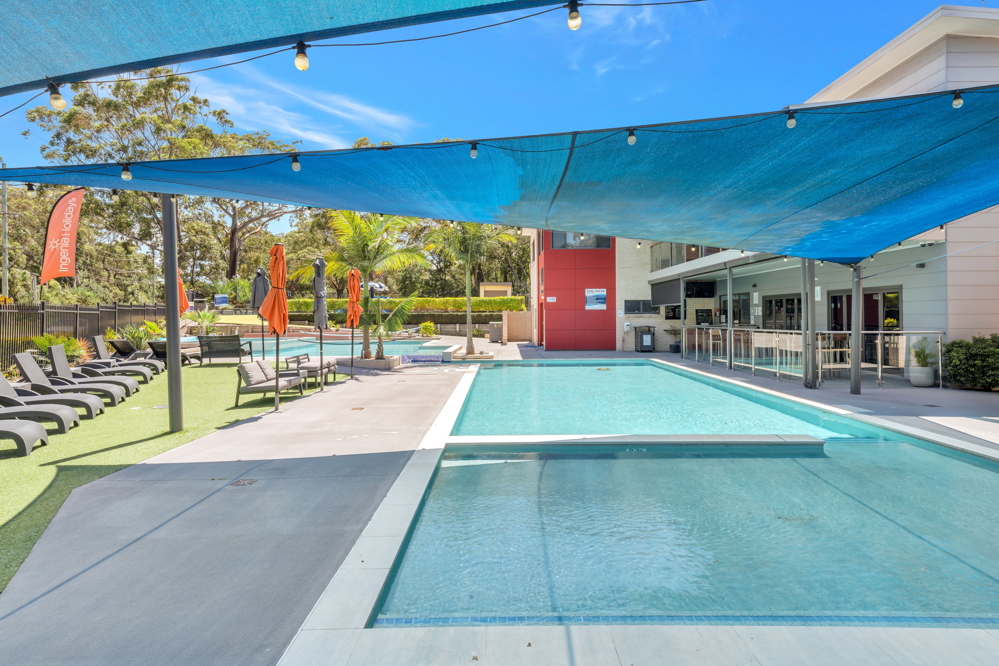 A swimming pool with lounge chairs and umbrellas, surrounded by a shaded area and a building with a red facade.