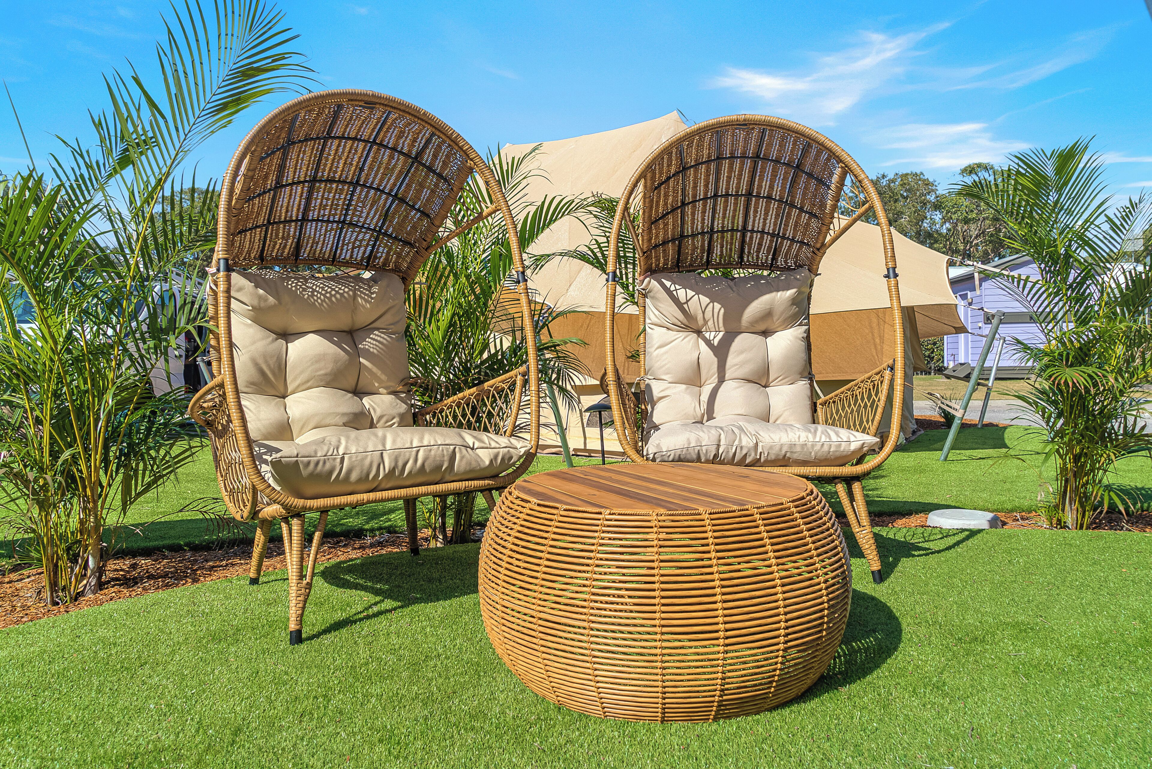 Two wicker chairs with cushions, a round wicker ottoman, and a beige canopy in a garden setting.