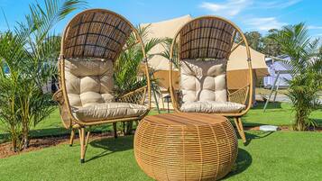 Twee rieten stoelen met kussens, een rond rieten pouf en een beige zonnescherm in een tuin.