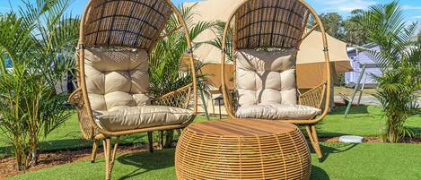 Two wicker chairs with cushions, a round wicker ottoman, and a beige canopy in a garden setting.