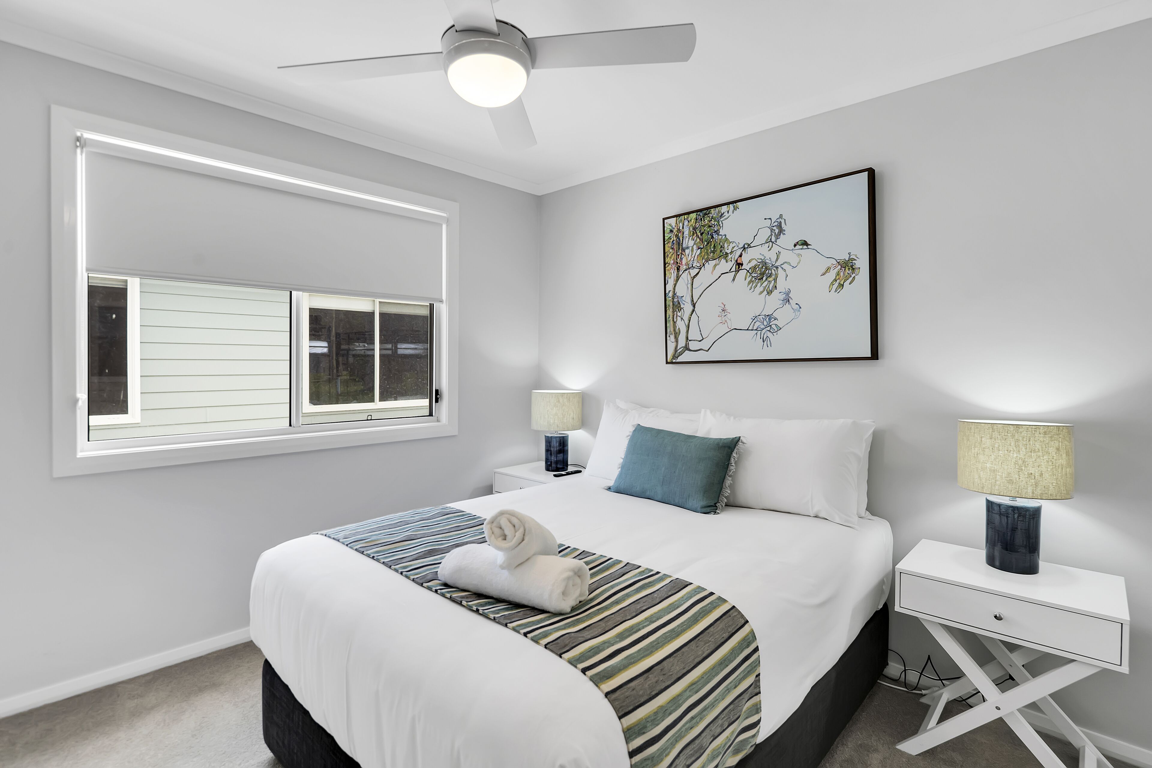 Une chambre moderne avec un grand lit, deux tables de chevet, un ventilateur de plafond et une fenêtre avec des stores.