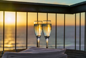Two champagne flutes on a wooden table with a white napkin, set against a sunset view through a balcony railing.