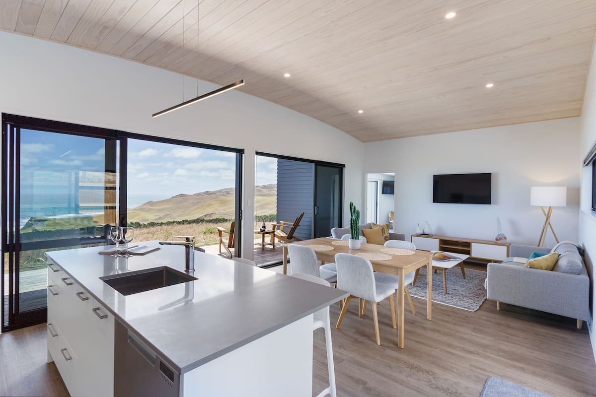 A modern kitchen with a large island, dining area with chairs, a sofa, and a TV mounted on the wall.