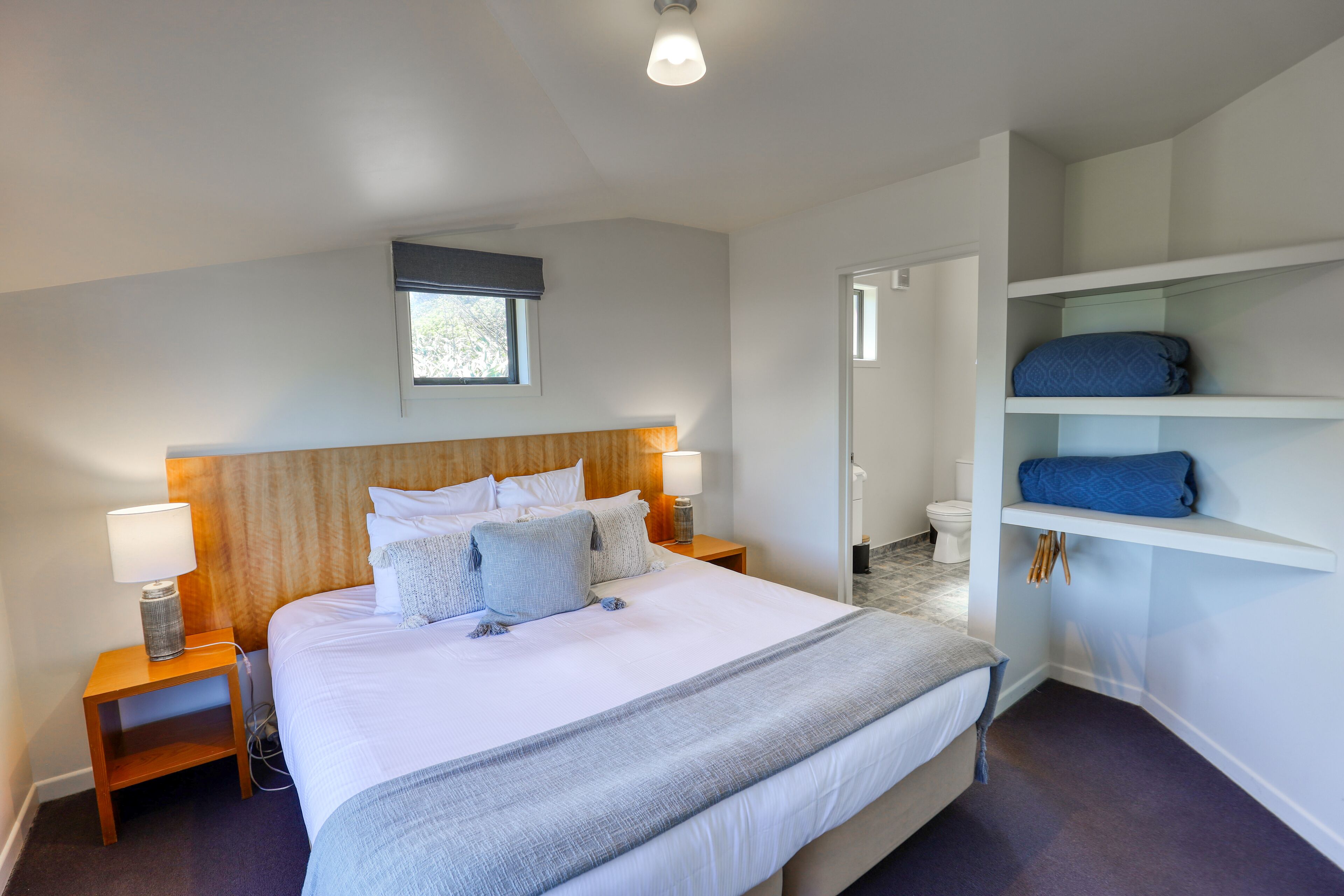 A modern hotel room with a large bed, bedside tables with lamps, a wooden headboard, a small window, and built-in shelving with folded towels.