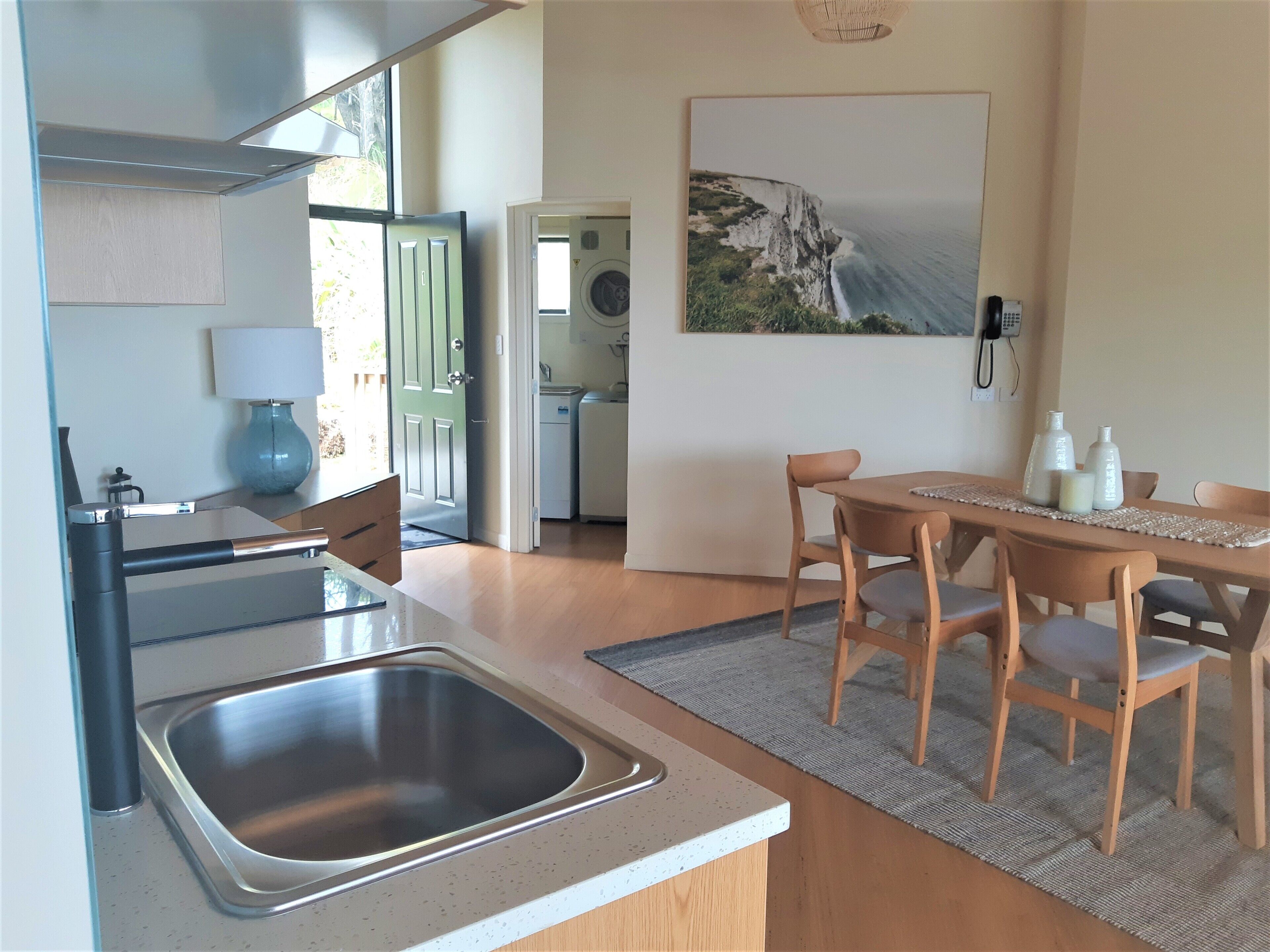 A modern kitchen with a stainless steel sink, wooden dining table, and chairs. There is a painting on the wall and a washing machine in the background.
