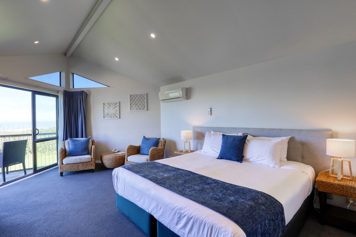 A modern hotel room with a large bed, two armchairs, a balcony with a view, and a ceiling with skylights.