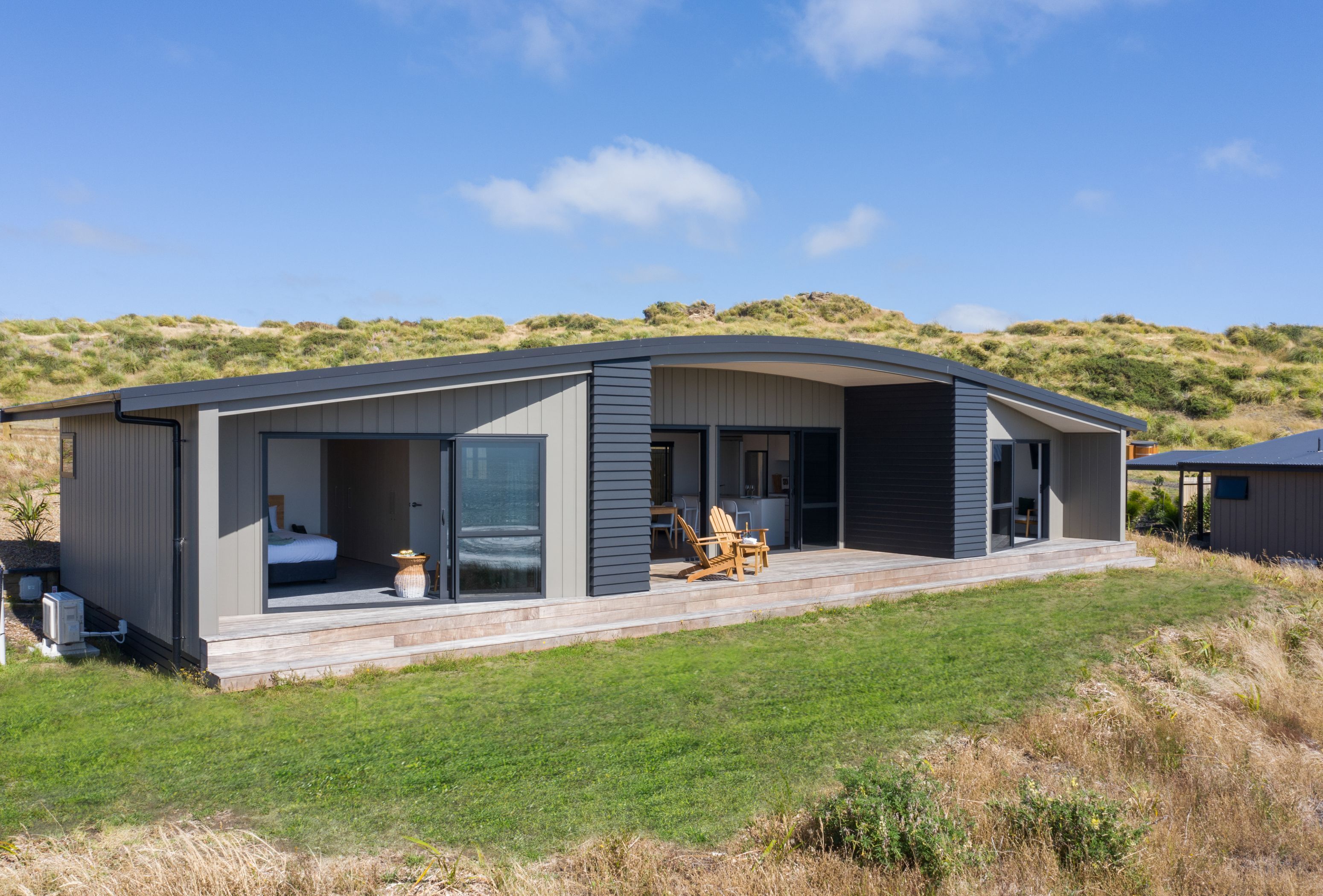 Une maison moderne dotée d’un grand patio, d’une chambre avec un lit et un fauteuil, et d’une cuisine visible à travers la porte ouverte.