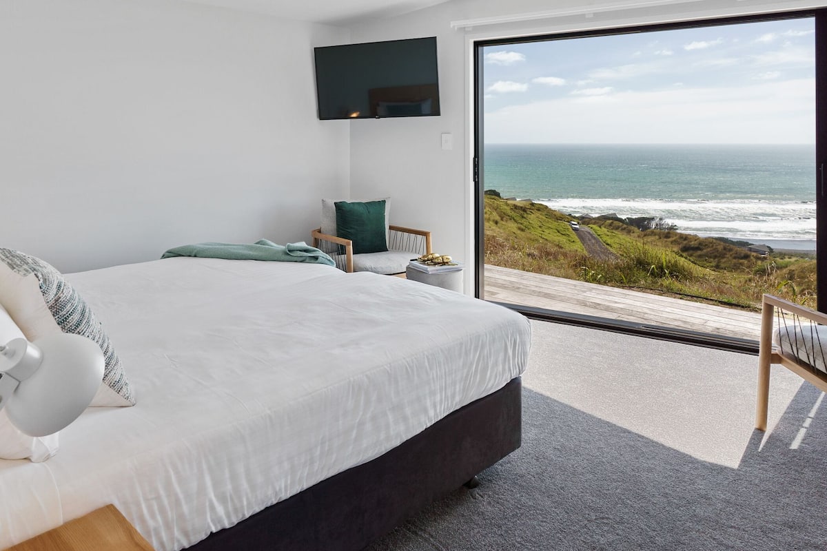 A bedroom with a large bed, a chair, a TV mounted on the wall, and a view of the ocean through a sliding glass door.