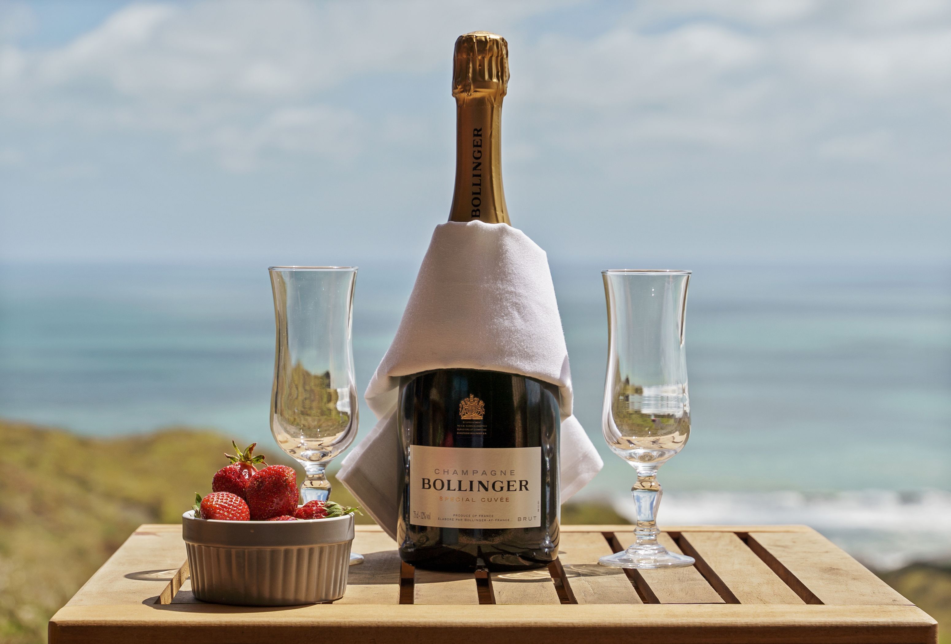 A bottle of Champagne Bollinger Special Cuvee on a wooden table with two glasses and a bowl of strawberries.