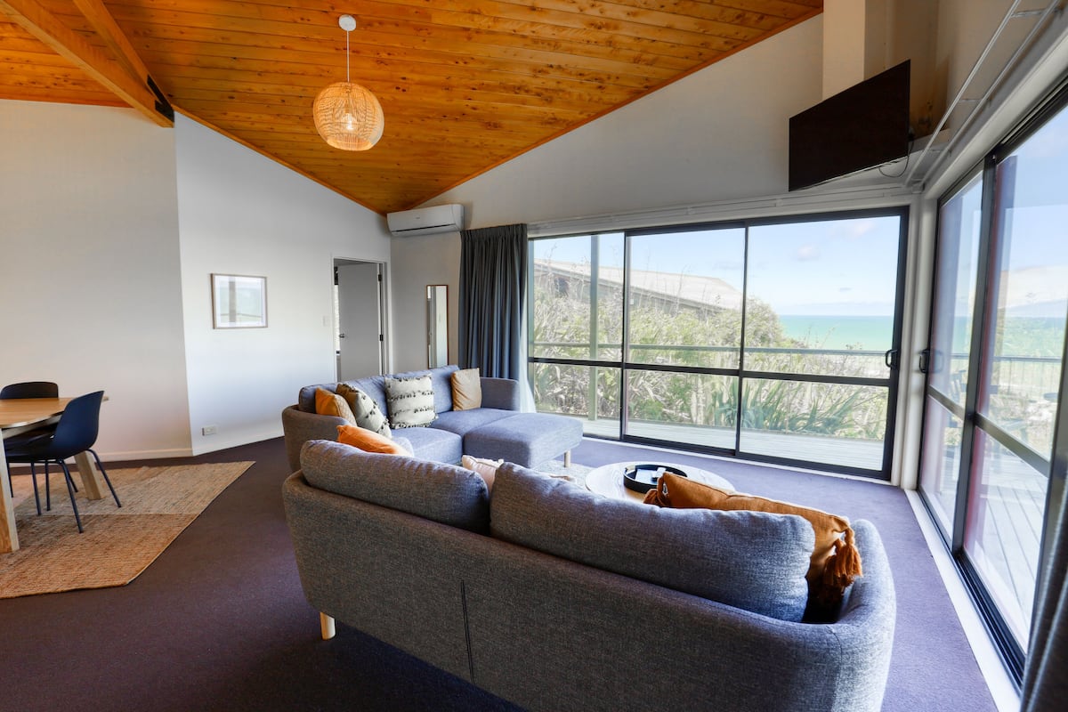 A modern living room with a large gray sectional sofa, a dining area with a table and chairs, and a large window offering a view of the outdoors.