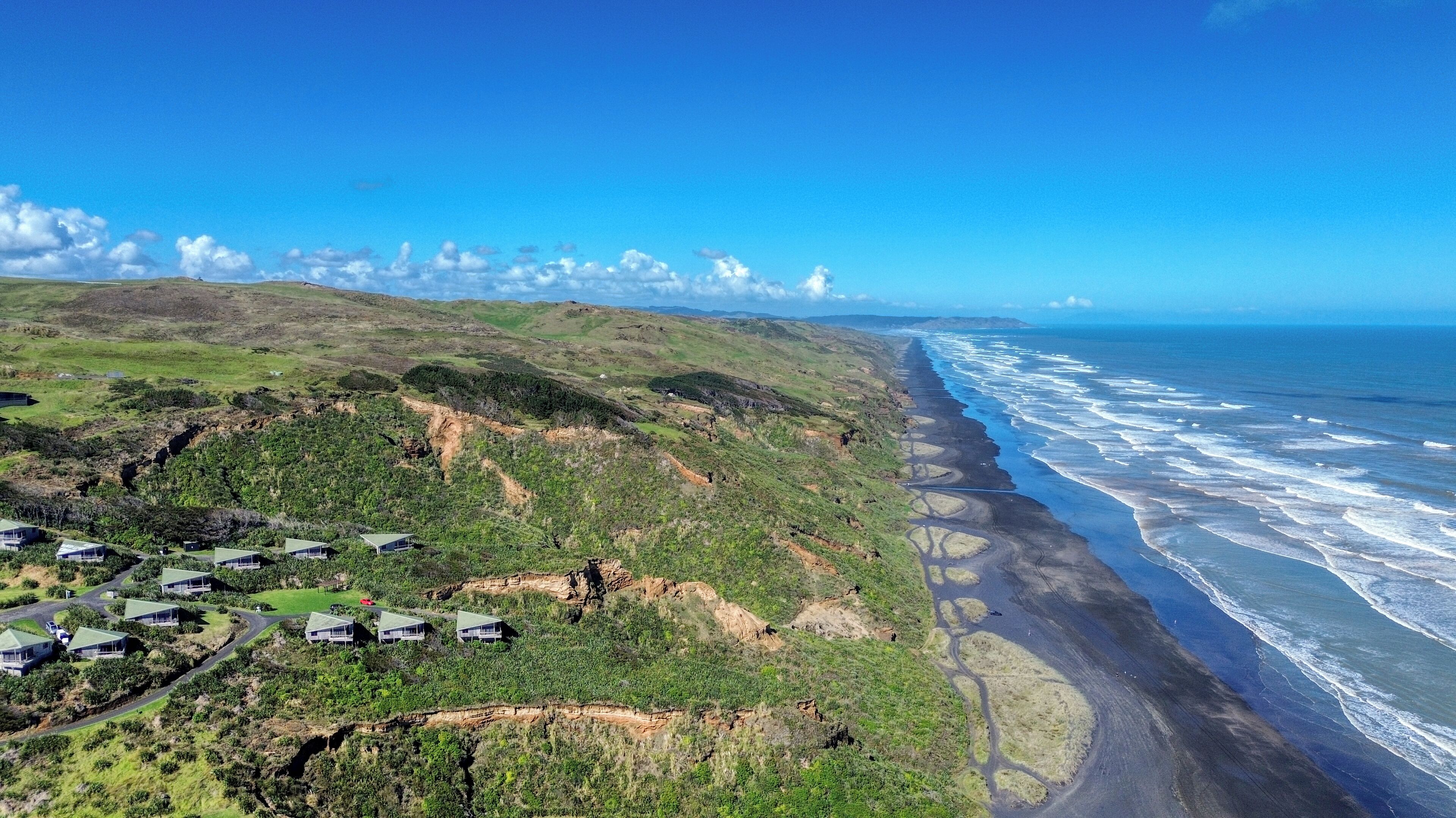 Un paisaje costero con una serie de edificios modernos de baja altura y techos planos, ubicados en una ladera con vistas a una playa donde las olas rompen contra la orilla.