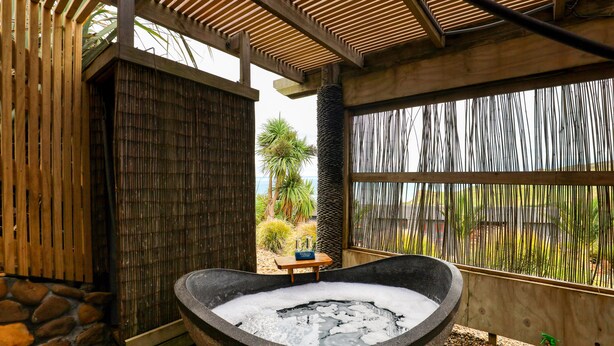 A stone hot tub with a wooden bench and a small table, surrounded by a bamboo fence and a stone wall. - Castaways Resort (Lake Puketi)