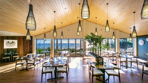 A spacious dining area with wooden tables and chairs, large windows offering a view of the ocean, and hanging pendant lights. - Castaways Resort (Lake Puketi)
