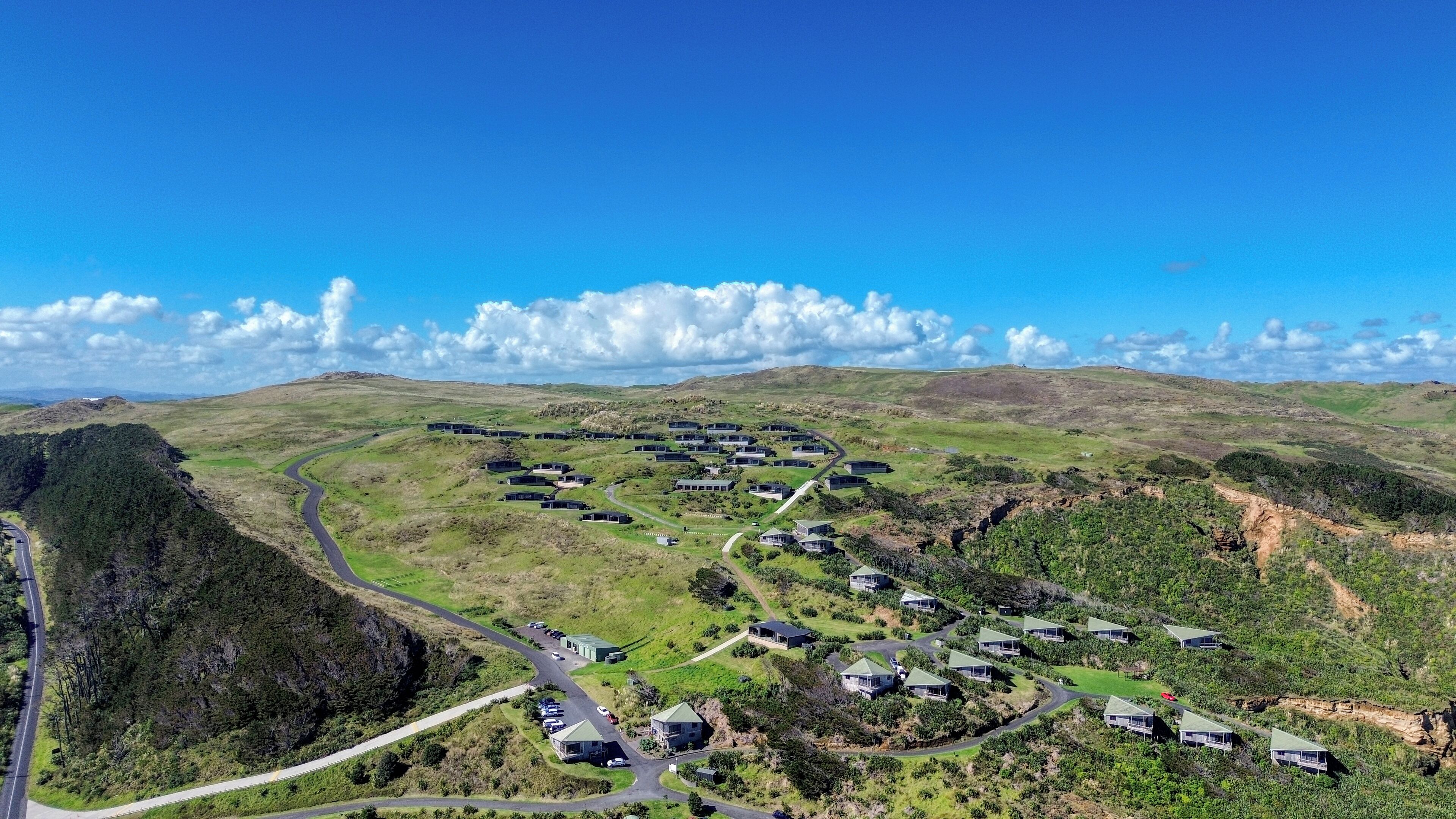 An aerial view of a residential area with houses arranged in a semi-circular pattern around a central road, surrounded by green hills and sparse vegetation.