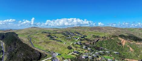 An aerial view of a residential area with houses arranged in a semi-circular pattern around a central road, surrounded by green hills and sparse vegetation.