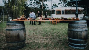 A rustic outdoor dining setup with a wooden table and barrels, surrounded by greenery and a white building in the background. - Karribank (Porongurup)