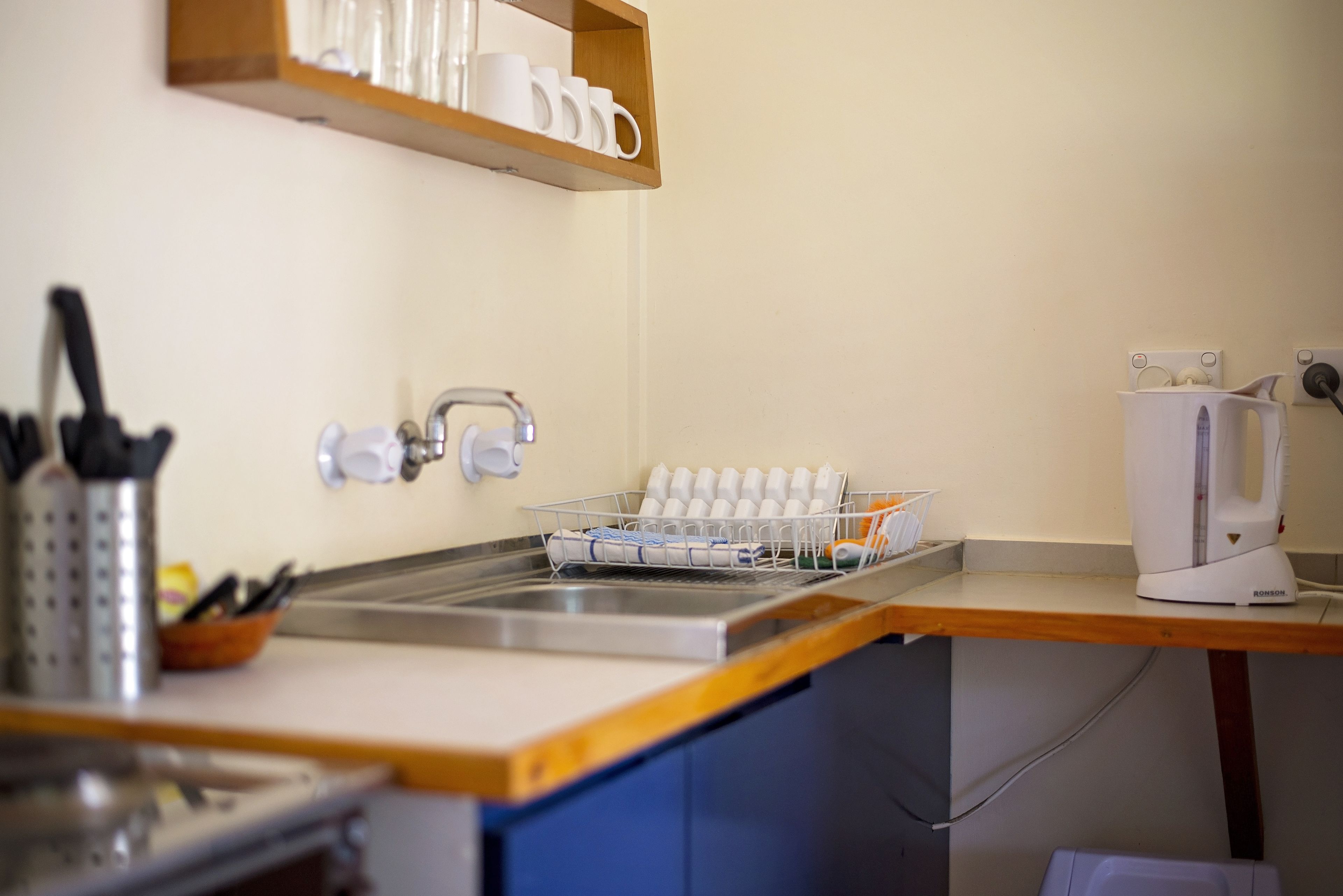 A kitchen with a sink, a dish rack, a coffee maker, and a shelf with cups.