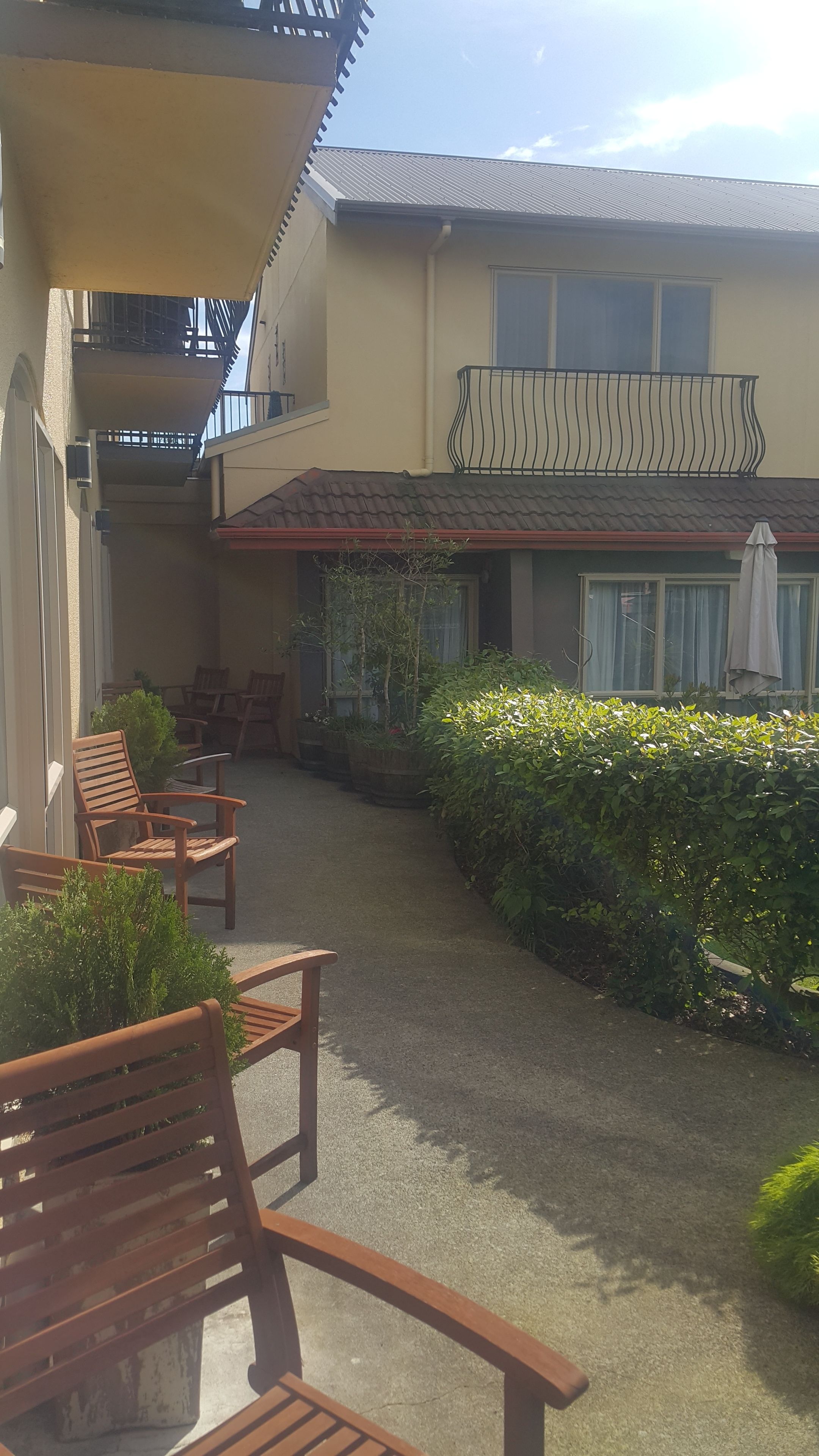 A courtyard with wooden chairs and potted plants, a building with a balcony, and a window with a curtain.
