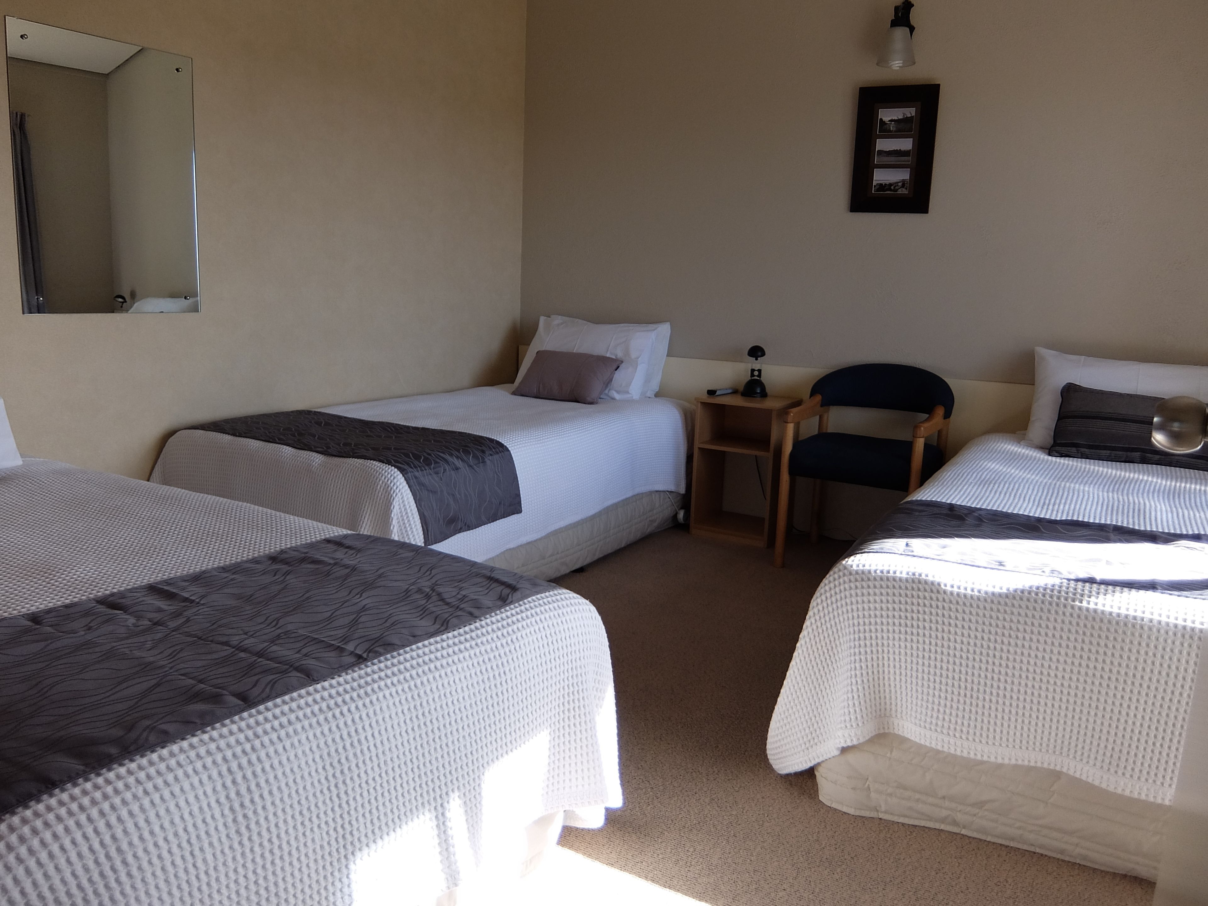 Habitación de hotel con tres camas individuales, una mesita de noche de madera, una silla, un espejo y un cuadro en la pared.