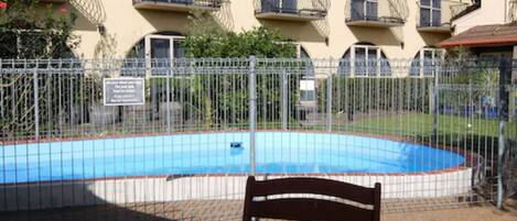 A pool area with a wooden bench, surrounded by a metal fence, and a building with balconies in the background.