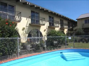 Una piscina con valla metálica, un edificio con balcones y un cielo azul despejado.