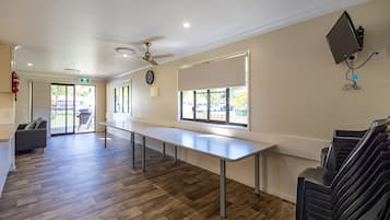 Una habitación amplia con una mesa larga, un ventilador de techo, un reloj y una ventana con persianas.